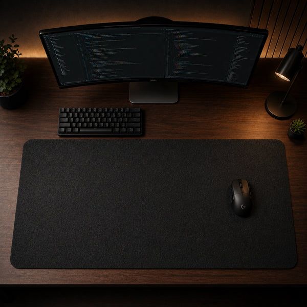 A large black desk mat on a wooden desk, styled with a black keyboard, a curved computer monitor, a black mouse, and a small desk lamp.