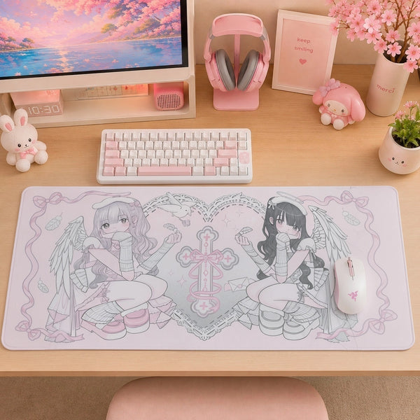 A large desk mat with a pink and gray anime-style illustration of two angel characters sits on a light wood desk. The desk is styled with a white and pink keyboard, pink headphones, a monitor, a small bunny figurine, and a pink vase with cherry blossoms.