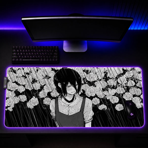 A large rectangular gaming desk mat with a black-and-white anime character illustration, shown on a desk under a monitor with blue LED lighting. The desk mat has a purple glow around the edges and includes a small logo in the bottom-right corner.